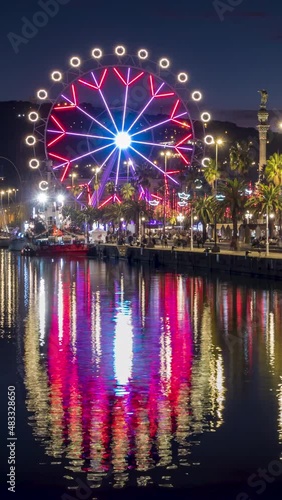 Cityscape with reflections in the water of the port of Barcelona,Catalonia,Spain. Ferris wheel rotating at dusk with many colorful led lights in motion with long exposure.Video in vertical format.