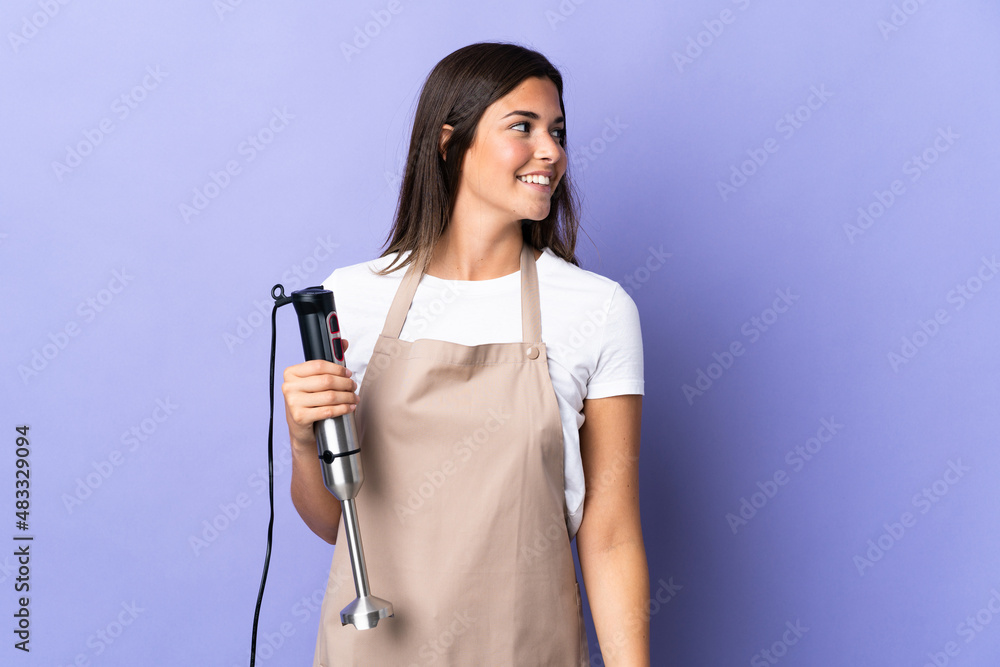 Brazilian woman using hand blender isolated on purple background looking to the side and smiling