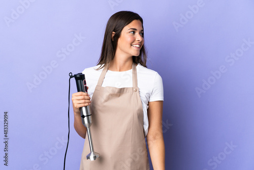Brazilian woman using hand blender isolated on purple background looking to the side and smiling