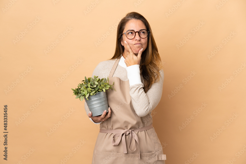 Middle age gardener woman holding a plant isolated on beige background having doubts Middle age gardener woman holding a plant isolated on beige background having doubts
