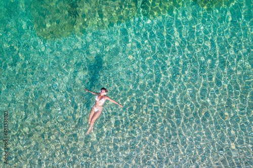 Top  aerial view. Athletic body of young beautiful woman in headphones listening music lying and swimming in white bikini in sea water on the sand beach. Drone  copter photo. Summer vacation.