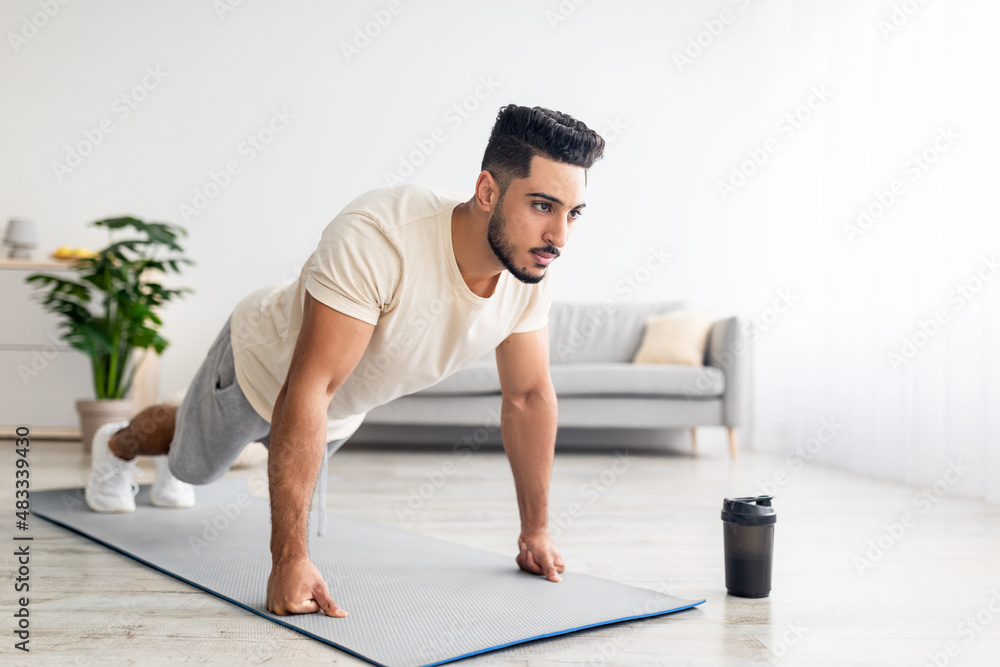 Domestic sports. Athletic young Arab man standing in plank pose, doing push ups, working out core muscles at home