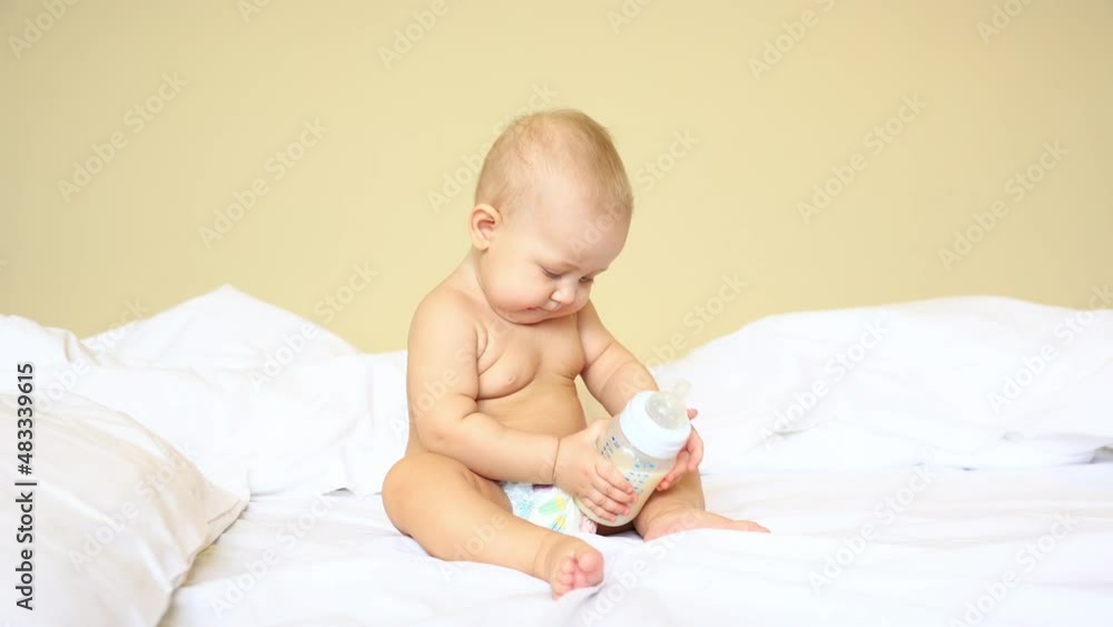 baby in a diaper is sitting on the bed in the bedroom hold bottle with milk mixture baby in a diaper is sitting on the bed in the bedroom hold bottle with milk mixture