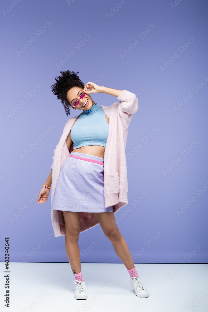 full length of positive african american woman adjusting trendy sunglasses on violet. full length of positive african american woman adjusting trendy sunglasses on violet.