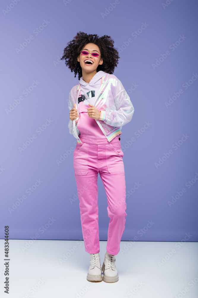full length of happy african american woman in pink sunglasses and overalls adjusting trendy jacket on purple. full length of happy african american woman in pink sunglasses and overalls adjusting trendy jacket on purple.