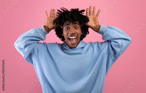 Cool African American teen guy making silly gesture with his hands, grimacing and having fun on pink studio background