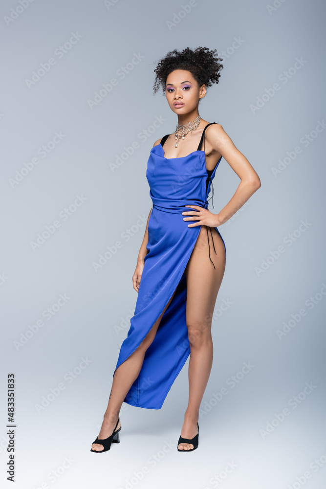 full length of african american woman in bright blue dress posing with hand on hip on grey.
