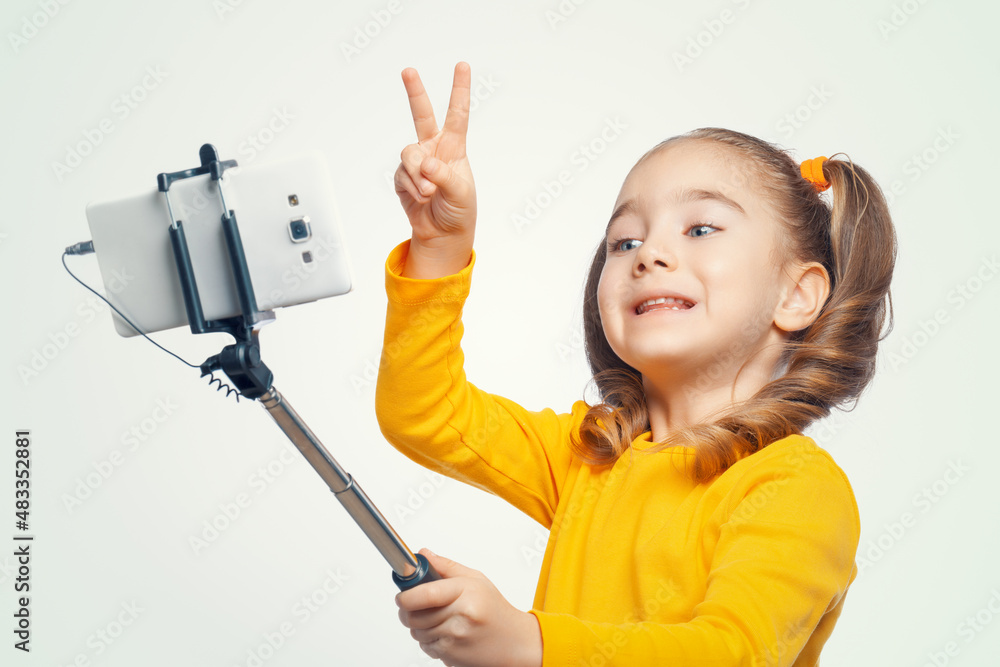 Portrait of smiling beautiful little girl holding selfie stick and making selfie on white background. Child making victory sign with her hand and posing to smartphone. Portrait of smiling beautiful little girl holding selfie stick and making selfie on white background. Child making victory sign with her hand and posing to smartphone.
