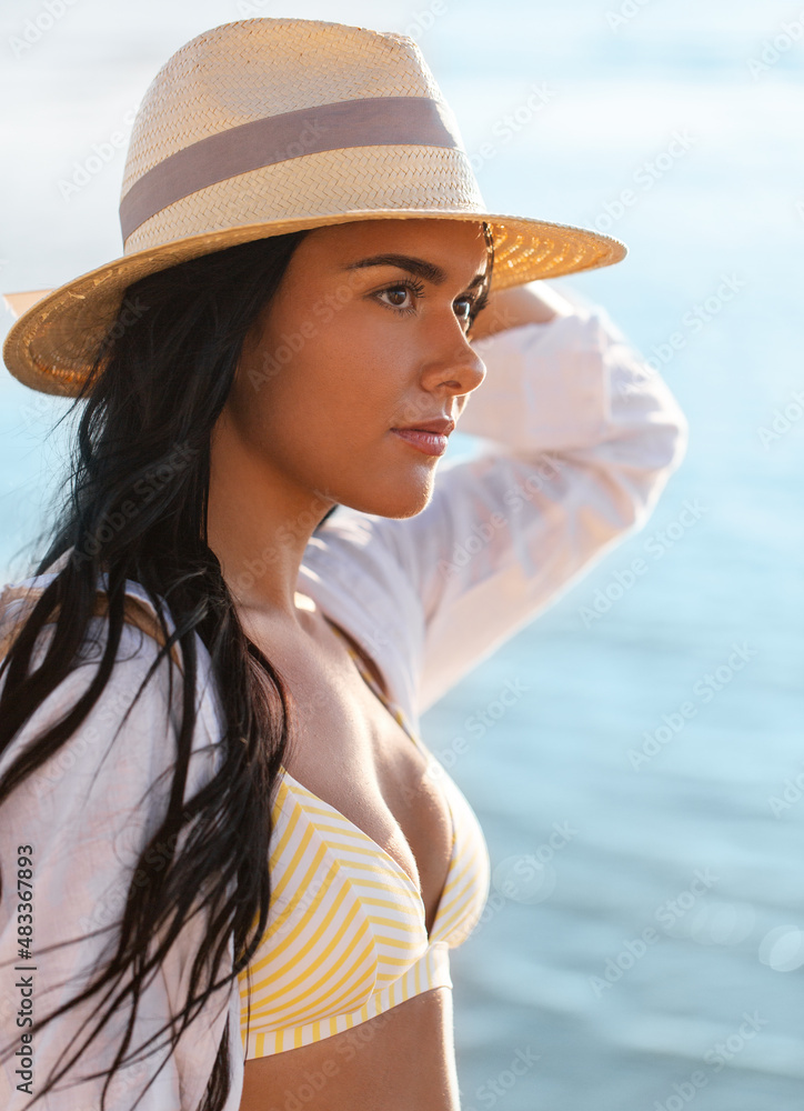 people, summer and leisure concept - portrait of young woman in bikini swimsuit, white shirt and straw hat on beach