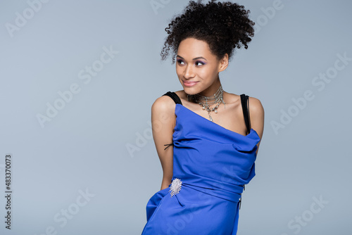 smiling african american model in slip dress looking away while posing isolated on grey.