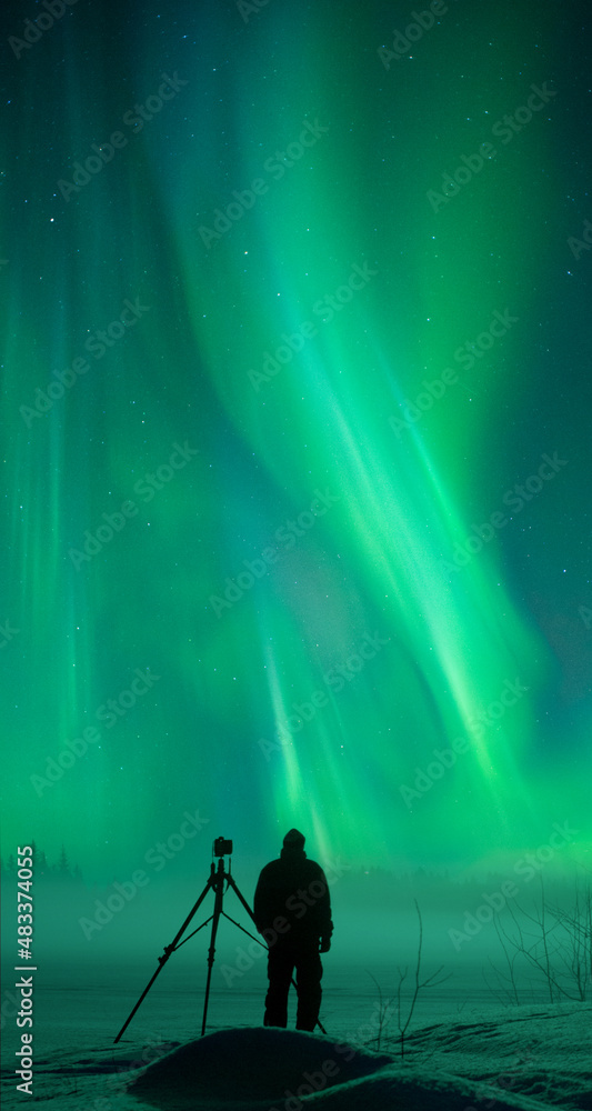 Photographer with his camera and tripod taking pictures of Northern Lights. Aurora Borealis over snowy winter landscape.