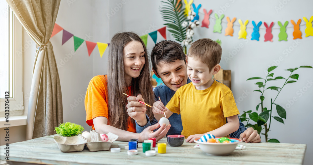 Fototapeta premium Mom, dad and little son in bright clothes are coloring eggs in a decorated room. Concept of family preparation for Easter, festive spring mood