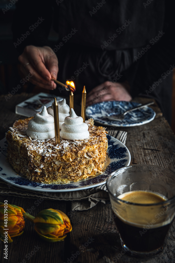 Woman lit candles on Mille-feuille cake decorated with meringues on vintage plate on old wooden table. Woman lit candles on Mille-feuille cake decorated with meringues on vintage plate on old wooden table.