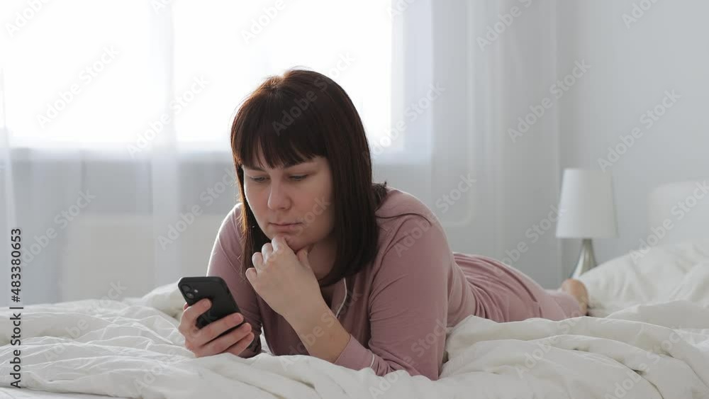 young woman in pink pajama lying on her bed and using smartphone in bright bedroom young woman in pink pajama lying on her bed and using smartphone in bright bedroom