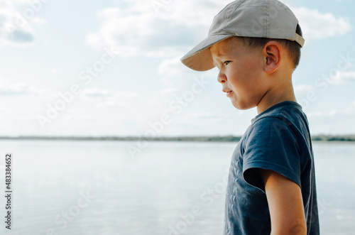 Portrait of sad little boy in cap standing alone by water on shore outdoors, copy space. Side view caucasian child in an unhappy mood in nature © Sergio