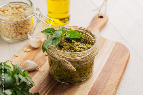 Jar of homemade green pesto sauce with fresh basil on a light wooden table. Mediterranean cuisine.