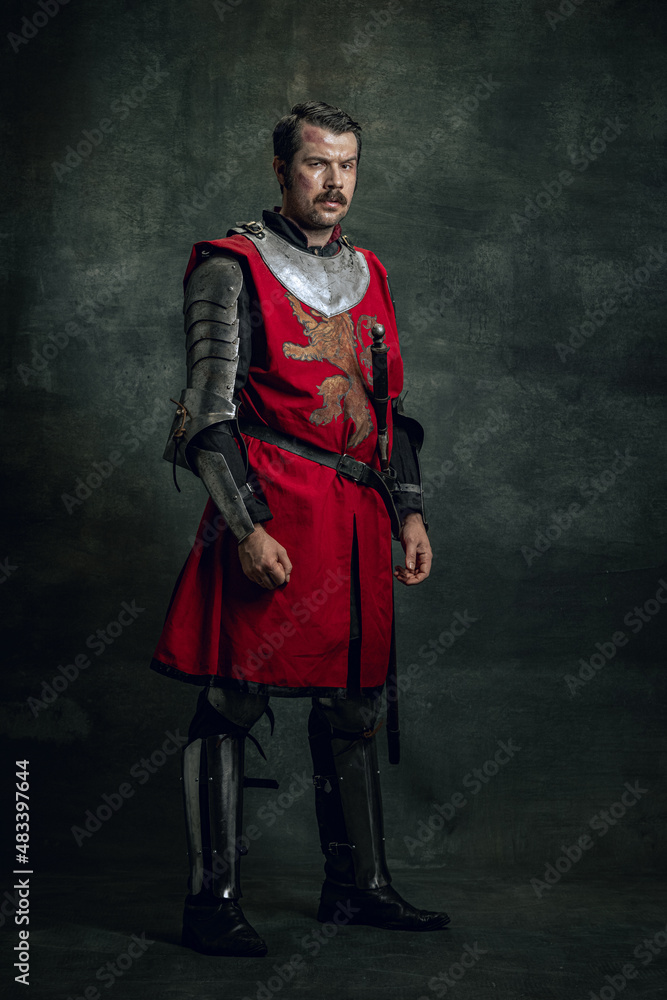 Vintage style portrait of brutal seriuos man, medieval warrior or knight with dirty wounded face holding sword isolated over dark background. Comparison of eras