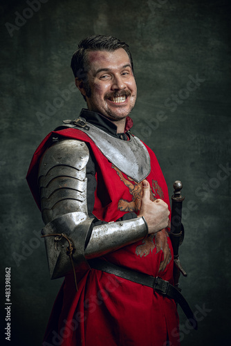 Portrait of smiling man in image of medieval warrior with dirty wounded face wearing equipment isolated over dark background. Comparison of eras
