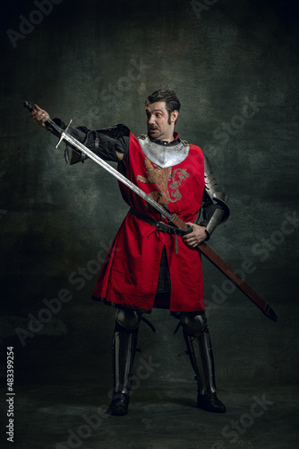 Comic portrait of funny medieval warrior or knight with dirty wounded face holding sword isolated over dark background. Comparison of eras