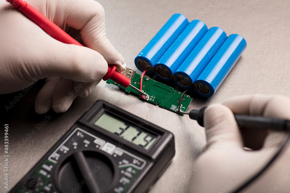 Charge indicator circuit, multimeter red and black poles in the hands of man checking voltage and amps, 18650 blue batteries, gray background. Powerbank
