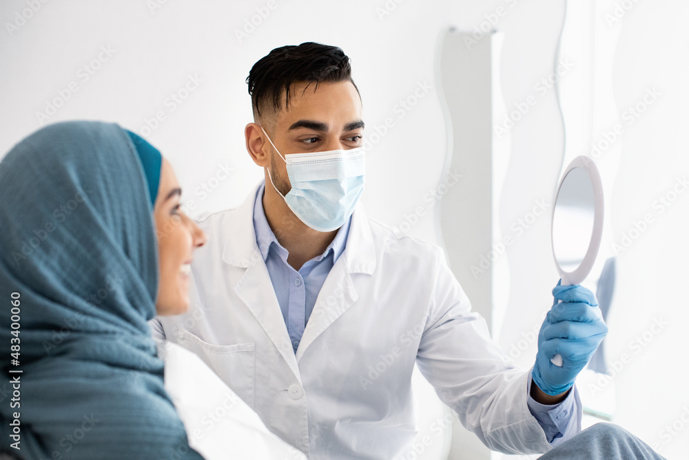Arab Stomatologist Holding Mirror, Showing Teeth Treatment Result To Muslim Lady Patient