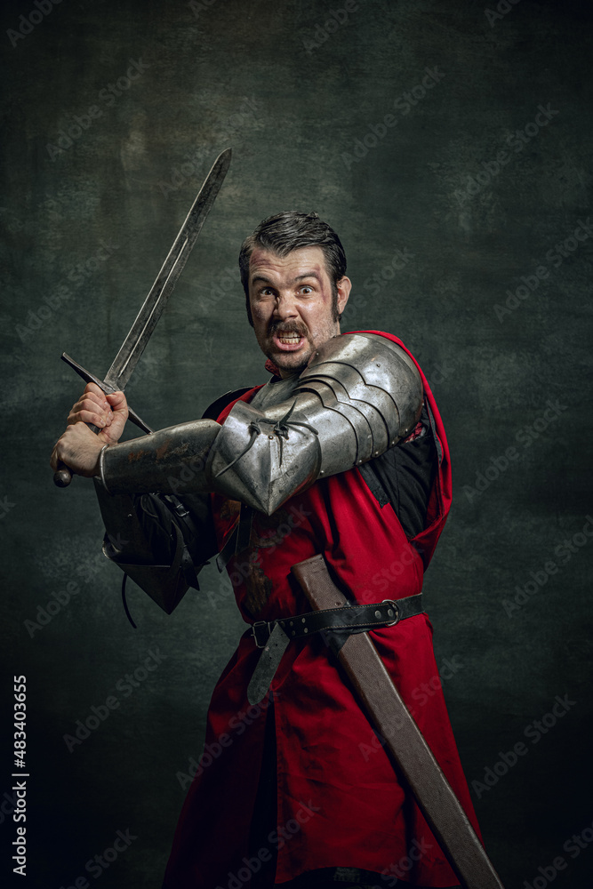 Dynamic portrait of seriuos man, medieval warrior or knight with dirty wounded face holding sword isolated over dark background. Comparison of eras