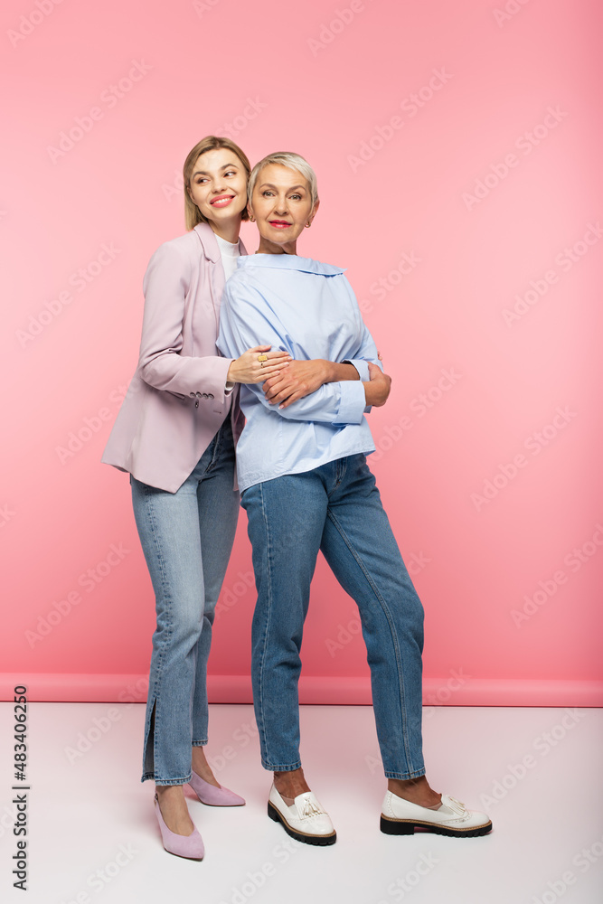 full length of happy young woman hugging mature mother in jeans and blouse while standing on pink full length of happy young woman hugging mature mother in jeans and blouse while standing on pink