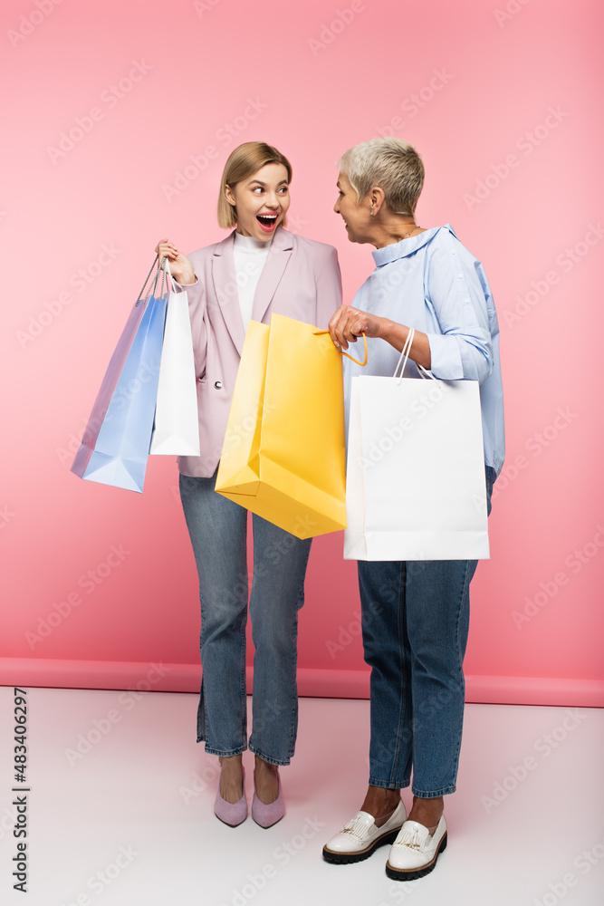full length of excited young woman and mature mother in jeans holding shopping bags on pink full length of excited young woman and mature mother in jeans holding shopping bags on pink