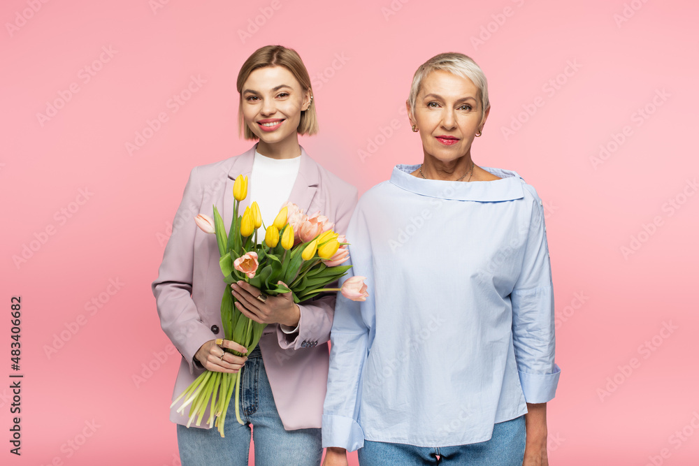 happy young daughter holding bouquet of tulips near middle aged mother isolated on pink