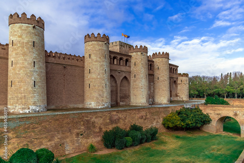Zaragoza, Spain - November 29, 2021: Medieval stone facade with towers of Aljaferia Palace in Saragossa - outside view. Ancient fortress, moat around with green grass and bushes and arch bridge