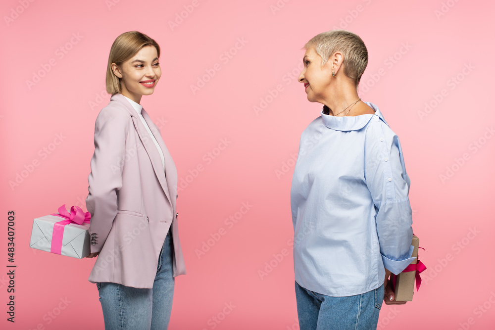 cheerful young daughter and mature mother holding presents behind back isolated on pink cheerful young daughter and mature mother holding presents behind back isolated on pink