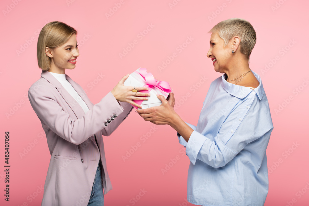 happy woman giving wrapped present to cheerful mature mother isolated on pink