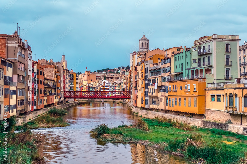 Panorama of Girona with red metal lattice Eiffel Bridge over the river Onyar, Spain. Catalan old town with colorful buildings on the shoreline of the stream and their reflections in the water