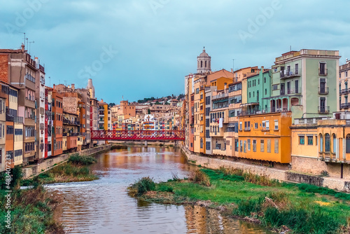 Panorama of Girona with red metal lattice Eiffel Bridge over the river Onyar, Spain. Catalan old town with colorful buildings on the shoreline of the stream and their reflections in the water © ioanna_alexa