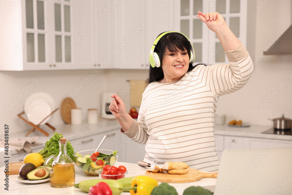 Happy overweight woman with headphones dancing while cooking in kitchen