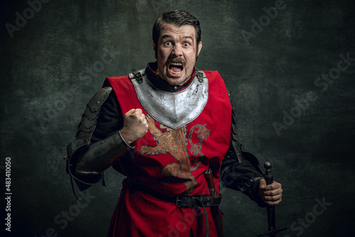 Half-length portrait of brutal seriuos man, medieval warrior or knight with dirty wounded face holding sword isolated over dark background. Comparison of eras