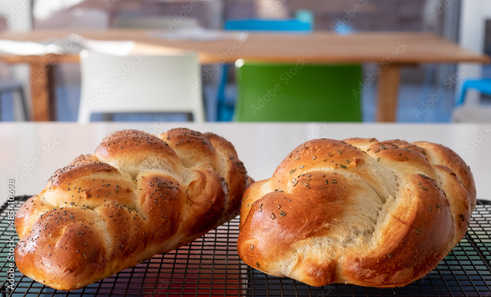 Two traditional home cooked plaited Jewish Challah bread loaves, cooling on a wire tray. One is topped with poppy seeds and the other with rosemary. Two traditional home cooked plaited Jewish Challah bread loaves, cooling on a wire tray. One is topped with poppy seeds and the other with rosemary.