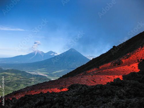 Volcanic river flowing between the solidified lava in the Pacaya volcano in Guatemala with an explosion of the Fuego volcano in the background with fumarole