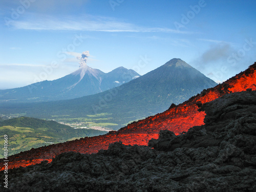 Volcanic river flowing between the solidified lava in the Pacaya volcano in Guatemala with an explosion of the Fuego volcano in the background with fumarole photo