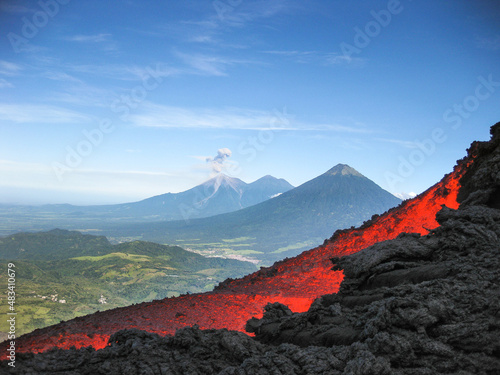 Volcanic river flowing between the solidified lava in the Pacaya volcano in Guatemala with an explosion of the Fuego volcano in the background with fumarole