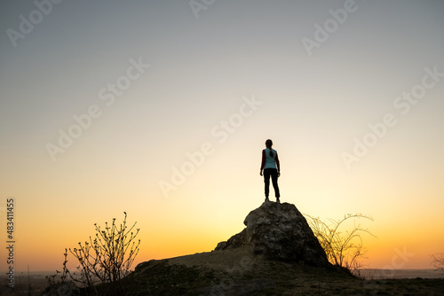Silhouette of a woman hiker standing alone on big stone at sunset in mountains. Female tourist on high rock in evening nature. Tourism  traveling and healthy lifestyle concept.