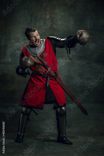 Portrait of medieval warrior or knight with dirty wounded face in boxing gloves isolated over dark background. Comparison of eras, history