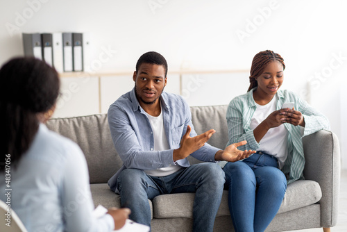 Fototapeta Naklejka Na Ścianę i Meble -  Irritated black guy complaining about his wife's smartphone addiction during psychological consultation at office
