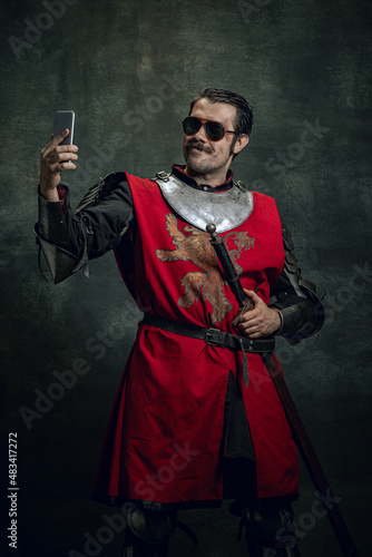 Smiling young man in image of medieval warrior or knight with dirty wounded face using phone isolated over dark background. Comparison of eras