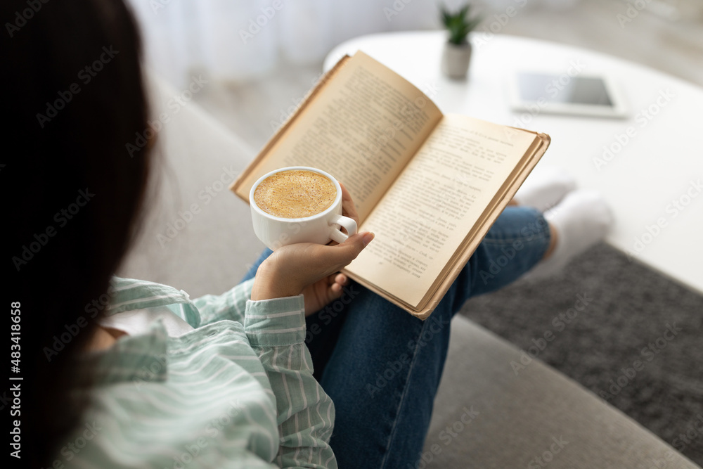 Naklejka premium Unrecognizable Indian woman reading book, drinking fesh cappuccino at home. Hobbies and pastimes concept