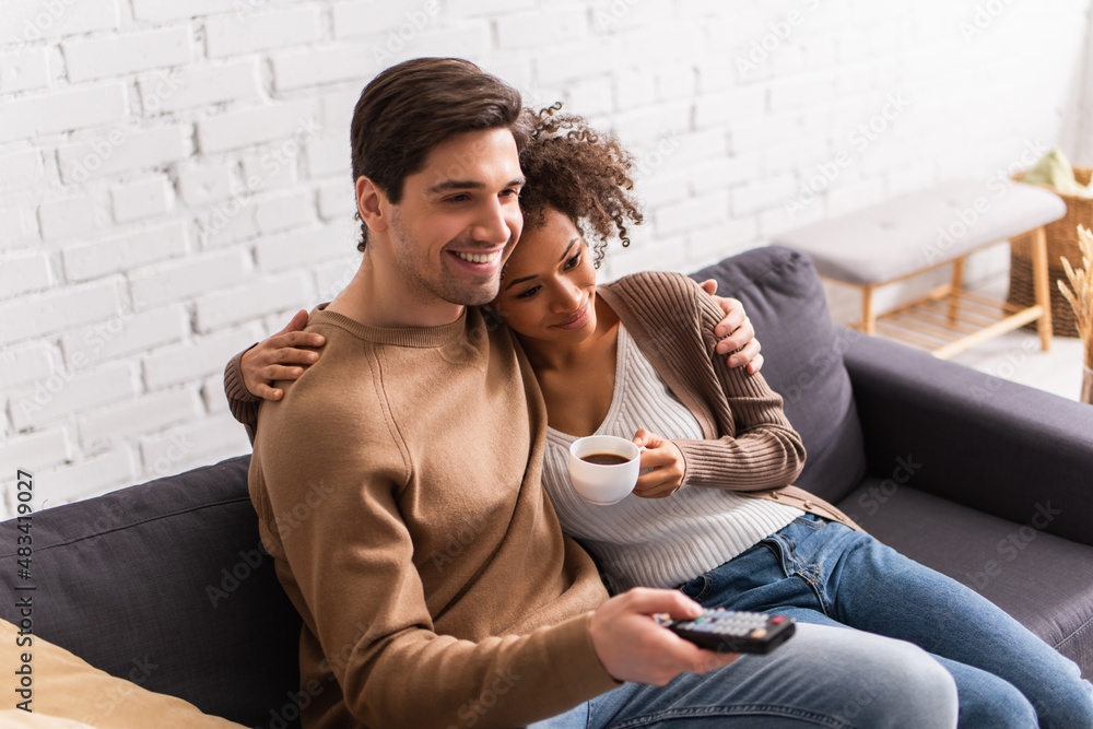 Smiling african american woman with coffee hugging boyfriend with remote controller at home Smiling african american woman with coffee hugging boyfriend with remote controller at home