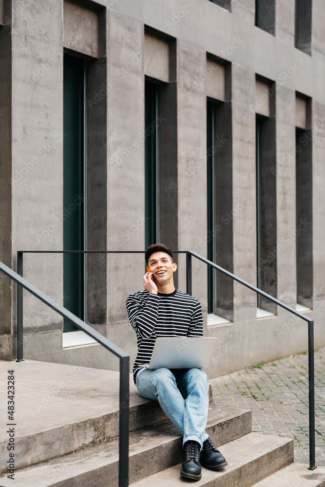 Young man, sitting on street stairs, using a laptop and talking on a red smartphone Young man, sitting on street stairs, using a laptop and talking on a red smartphone