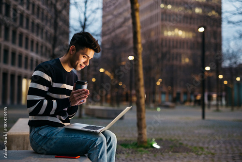 Young man, sitting on street bench, using a laptop and drinking coffee