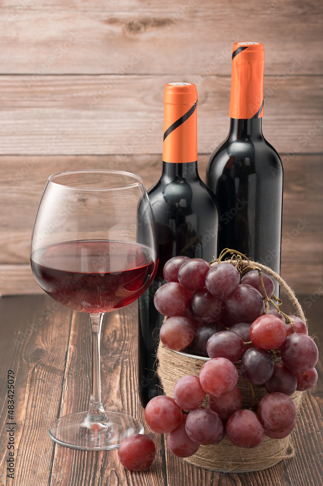 Fototapeta premium Glass of red wine with grapes on wooden table against the background of bottles