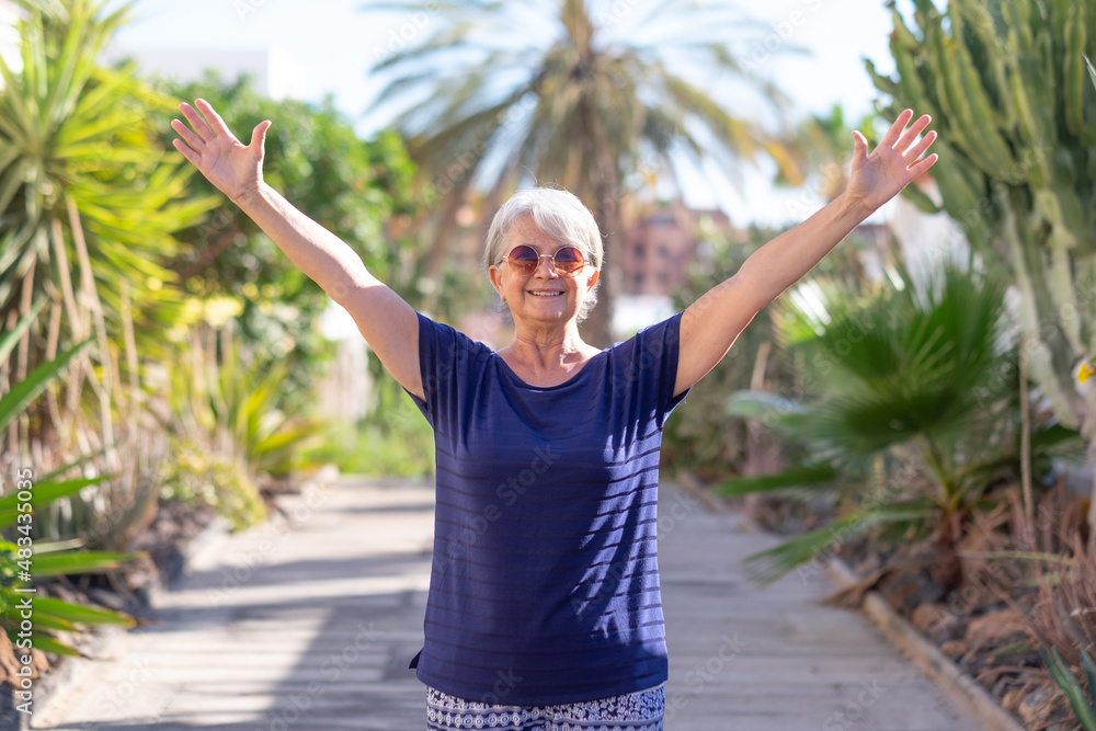 Happy smiling adult mature senior woman outdoor in a tropical garden with open arms. White haired caucasian people standing lin footpath enjoying sun, freedom and healthy lifestyle looking at camera